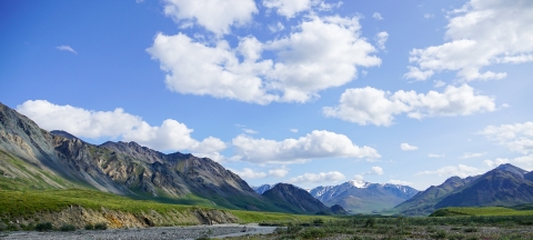 Landscape photo of green mountains, a river bank, and blue sky with clouds