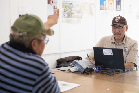 Two men at a table in discussion. One wears a Fish and Wildlife Service uniform. The other wears a cap and a striped shirt.