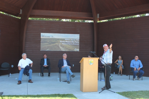 Demus McDonald guestures torwards the audience (out of frame) as he speaks. Behind him, seven people are seated.