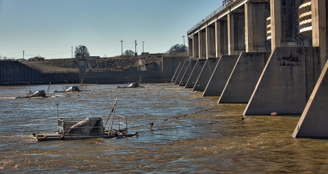 Four large pieces of metal equipment float in a river near a dam