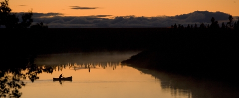 a person paddles a canoe across a river at sunset. 