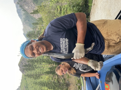 Photo of a smiling Black man with a lamprey stuck to his cheek, with a smiling Tribal woman behind him.