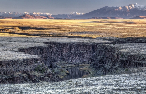 A river flowing through a canyon cutting through a snowy flat plain with snow-covered mountains in the distance