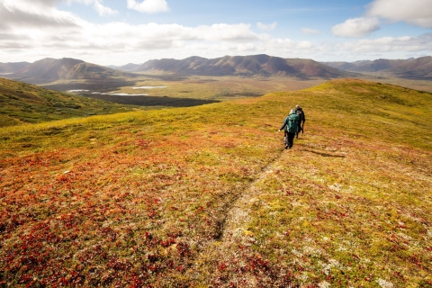 Two people with backpacks head out across a reddish plain with big mountain ranges in the distance.