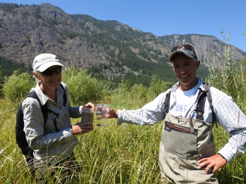 A woman and a man in waders and ballcaps stand in a field of waist-high plants with mountains in the background.