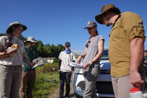 Five people standing outdoors in a circle as one points to a piece of paper