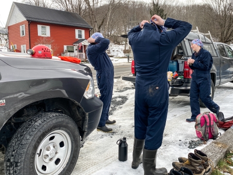 Three people standing beside a pair of trucks, putting on coveralls