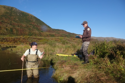 Volunteer in waders stands in a river holding a yard stick and water temperature probe while reporting results to a USFWS employee who records data on the bank.