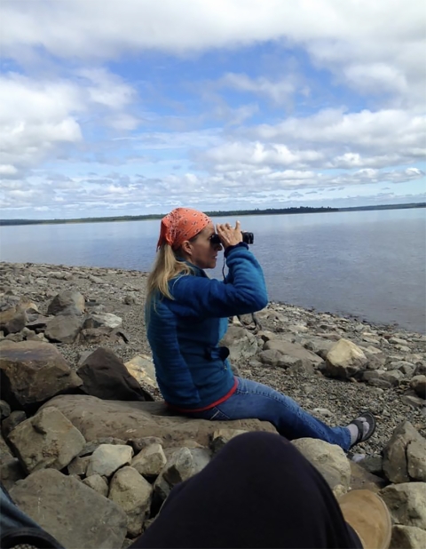 Person sits on rocks and looks over the water with binoculars.