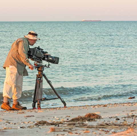 A man using a large video camera standing on a beach filming a crab in the water