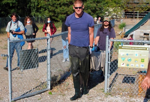 Two men carry a cage draped in a cloth out of a fenced area while other people watch.