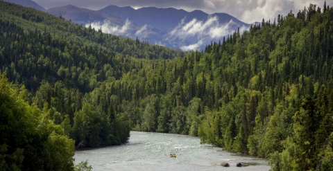 A boat heads down a river lined by forests and mountains.