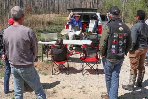 Several adults in a semi-circle, listening to one person sitting on a truck tailgate