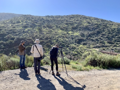 Three individuals standing on the trail looking towards a chaparral mountain. One person standing on the far right looks into a viewing scope.