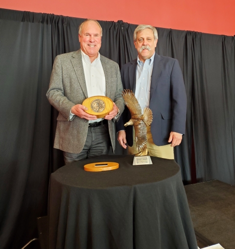 2 men behind table; one holds award