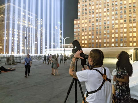 A person looks into a telescope aimed at the sky. A group of people stand on a rooftop looking up towards bright light beams.