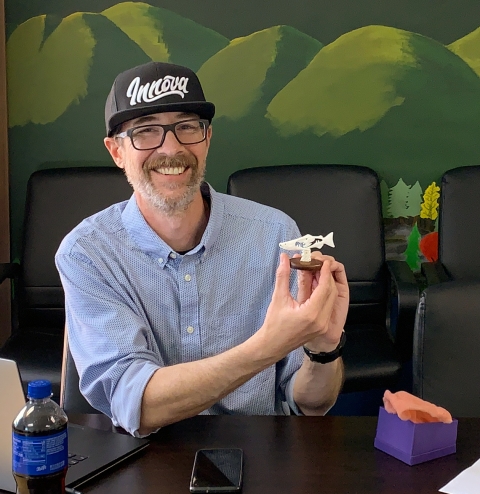 A smiling, bearded man in a ballcap holds up a small carved salmon