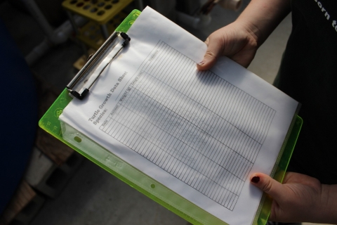 A student shows off a green clipboard with a paper. On the paper is a table labeled "turtle growth data sheet", partially filled in with pencil. 