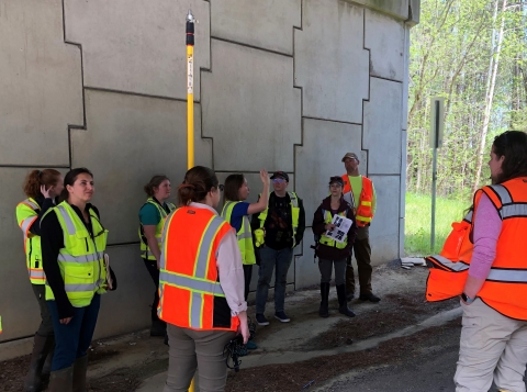 A group of people wearing brightly colored vests stand below a roadway bridge