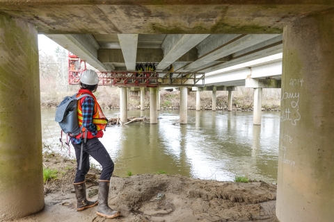 Person standing on a river bank beneath a bridge watching a crew working on a catwalk