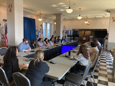 A group of 15 people sitting around a U-shaped conference table in a room with pillars and indigenous mural art decorating the walls.