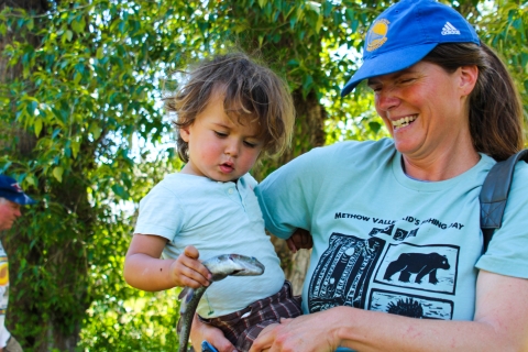 A young child held by a grinning woman examines a small trout.