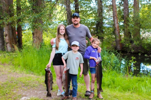 A man with two girls and a boy stand in front of trees, all 3 kids holding huge trout.