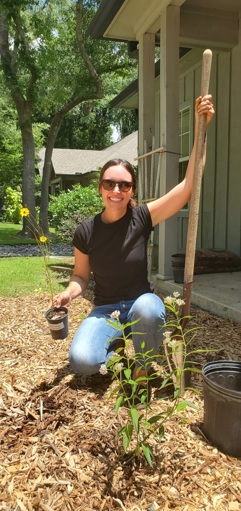 Jenna holding a shovel and a yellow flowered coreopsis plant posing for a picture before she plants the plant. She is kneeling behind a milkweed plant.