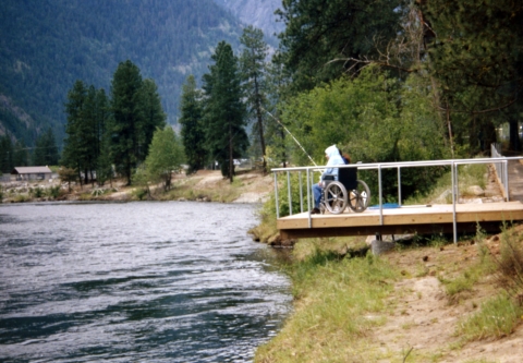 A man in a wheelchair and wearing a hoodie fishing from a platform overhanging a river.