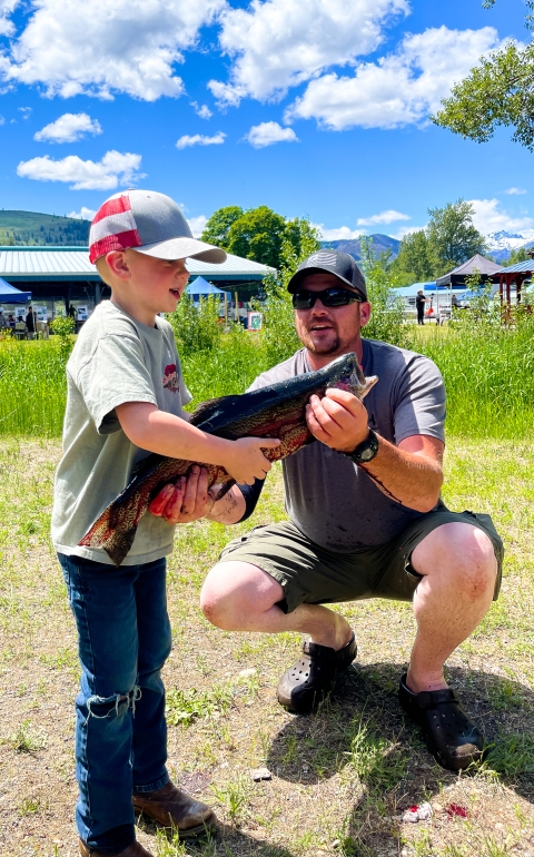 On a sunny day, a squatting man helps a boy support a large fish.