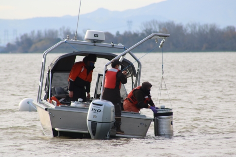 A woman on a boat lowers a large, cylindrical object into a river with assistance from a crane-like device