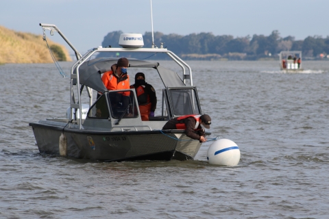 A man leans out of a boat on a river to attach a rope to a round, white buoy.