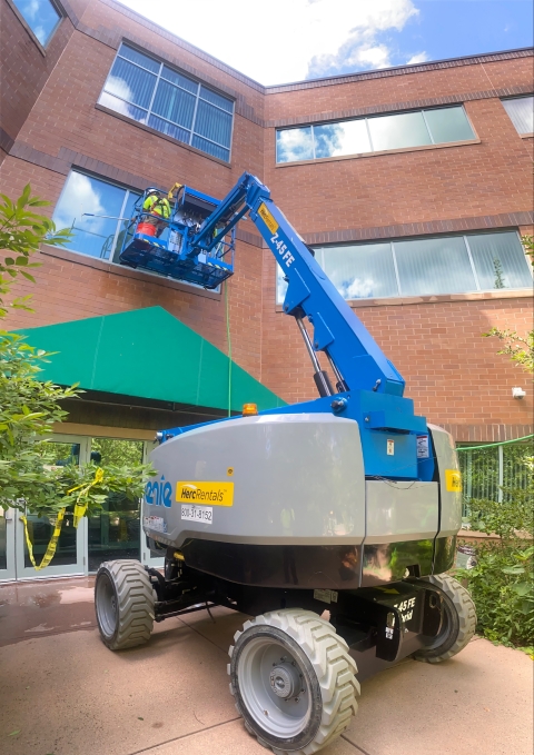 a man on a crane working on building windows. Clouds are reflected in the glass.