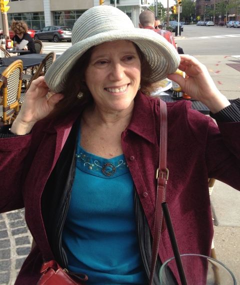 A woman in a blue blouse and maroon jacket holds a large straw hat on her head while sitting outdoors