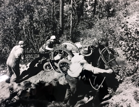 A black and white photo of a crew of 3 men urging a horse hauling a wheeled cart with a large piece of machinery in it up a steep, narrow, rocky trail.