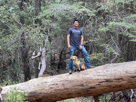 Man poses for a photo as he stands on downed tree trunk with dog sitting between his legs.