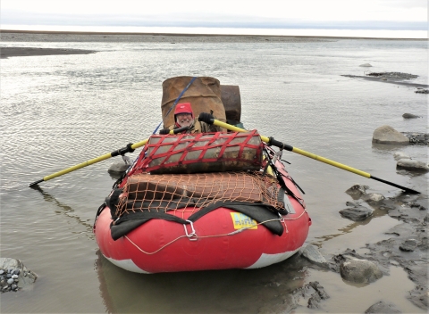 A man in a large red float boat with yellow oars in shallow water near shore