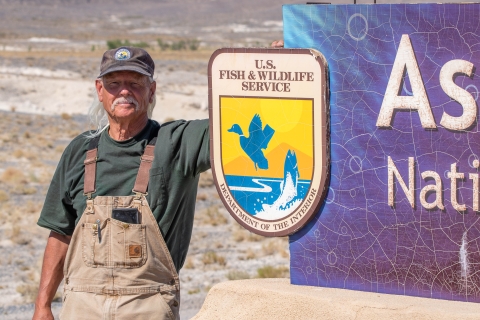 A man with a white mustache wearing overalls and a U.S. Fish and Wildlife Service volunteer cap standing next to a sign with the U.S. Fish and Wildlife Service logo on it