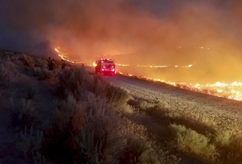 A fire truck driving down a dirt road with a fire on the right side of the road.