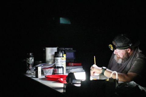 A bearded man sits in a dark environment. He has a cave lamp on his head. In front of him is a table filled with monitoring equipment. He writes in a notepad.