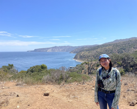 A woman smiling for the camera, standing in an open space overlooking the water