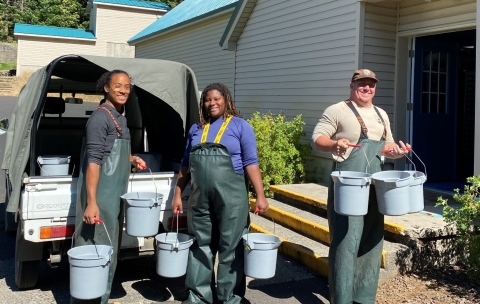 Two women and one man stand smiling in the sun in waders holding blue buckets.