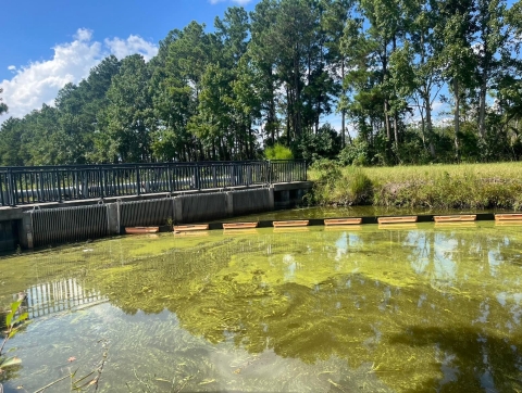 A concrete bridge structure with vertical grates along the sides extending into the water. In the foreground, a floating boom reaches diagonally across the canal.