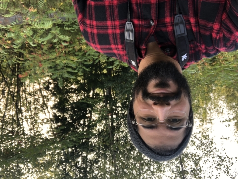 a bearded man with glasses and a beanie hat smiles while standing in a lush green forest. A black binocular strap hangs around his shoulders