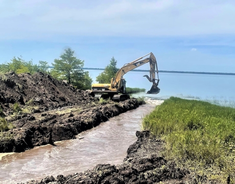 A bulldozer at the side of a large body of water digging a canal from the body of water