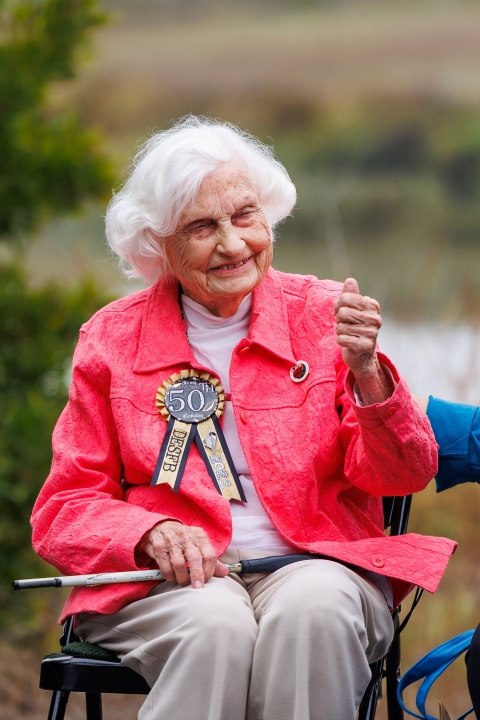 A woman wearing a gold pin reading "50th" smiles and gives a thumbs up