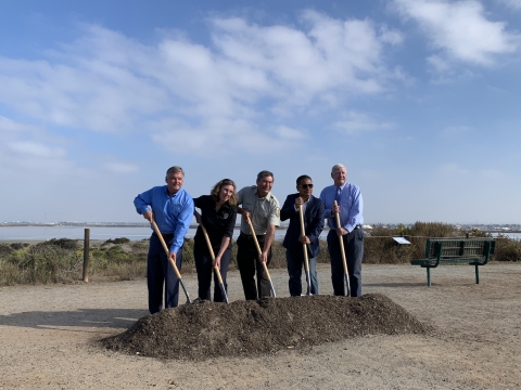 Five people with shovels get ready to dig into a pile of dirt with coastal habitat behind them.