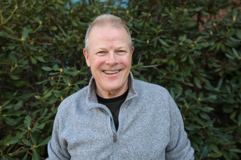 Headshot of a white man in a grey pullover against a leafy backdrop