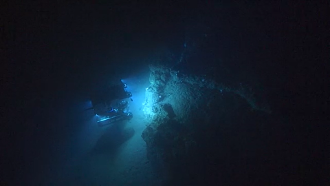 a submarine vessel shines lights on a rocky surface deep underwater. Darkness surrounds the vehicle