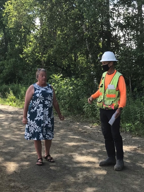 woman in blue dress talks to a person in a hard hat and reflective vest on a dirt path in the woods on a sunny green day 